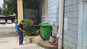 A person cleaning a tiled pavement area with a hose, a green trash bin, and a broom near a building with a blue door. - Feicui Hotel (Mangshi)