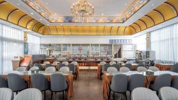 Un salón de conferencias con mesas y sillas dispuestas para reuniones, una gran lámpara de araña y vistas de la ciudad a través de amplios ventanales.