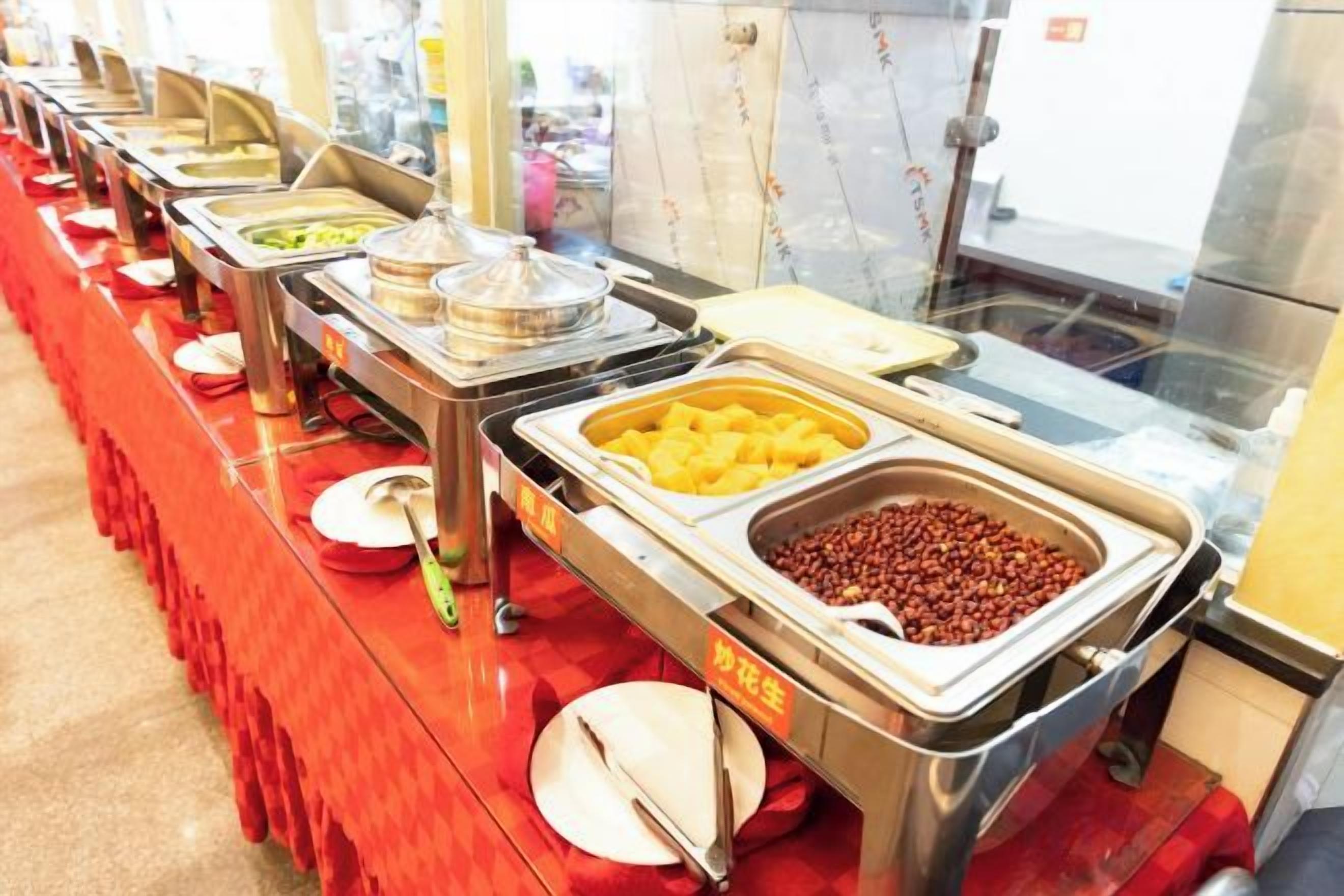 A buffet line with various food items in metal containers, a red tablecloth, and a glass partition.