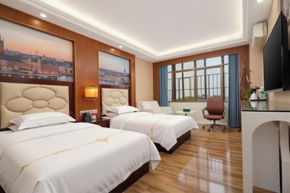 Quarto de hotel com duas camas, uma escrivaninha, uma cadeira, televisão e uma janela grande.