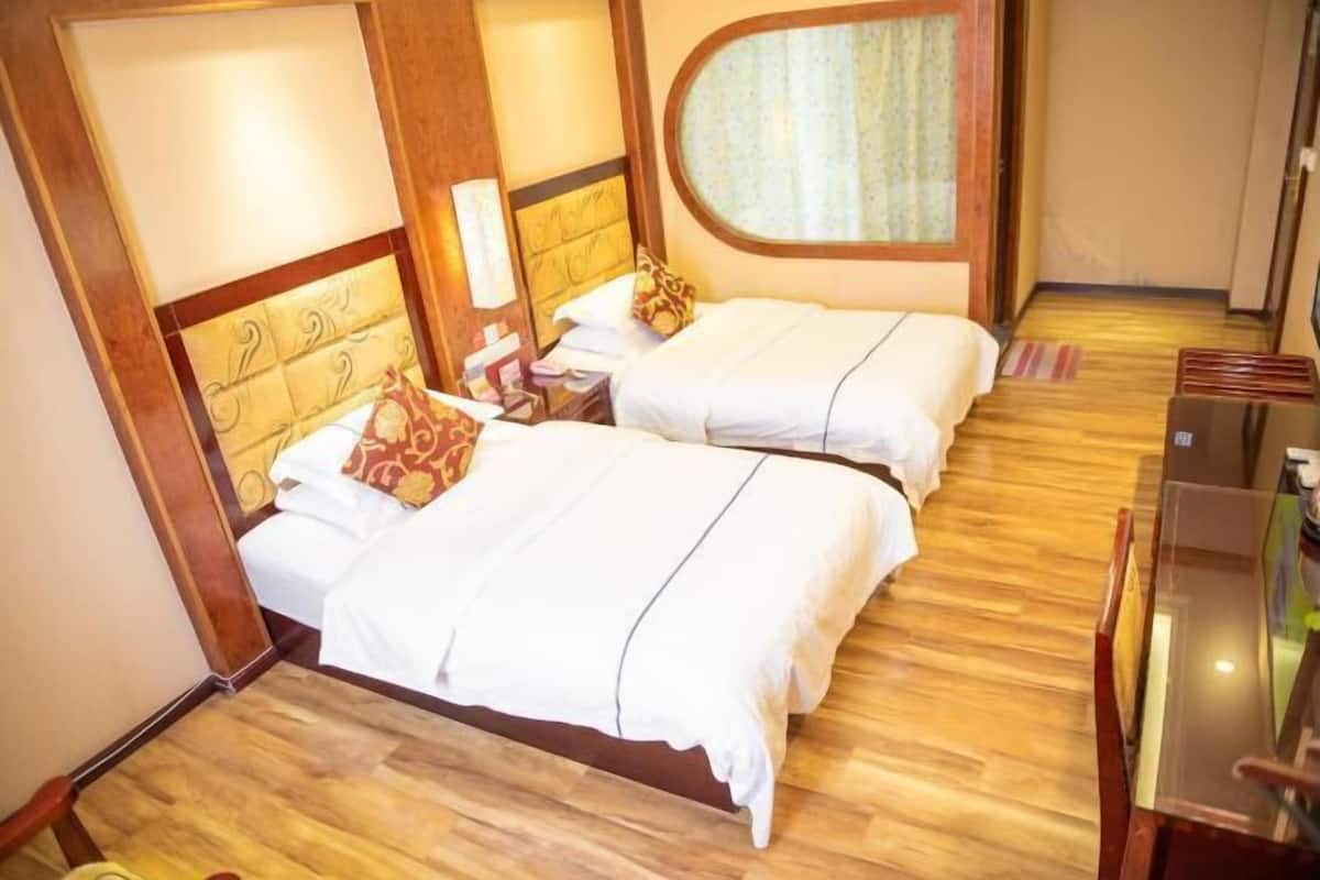 A hotel room with two beds, wooden flooring, a small desk, and a window with curtains.