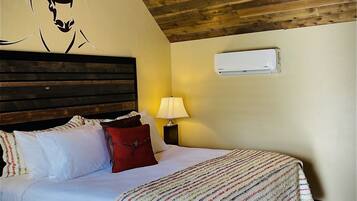 A bedroom with a wooden ceiling, a bed with white bedding and colorful striped blankets, a red pillow, and a wall-mounted air conditioner.
