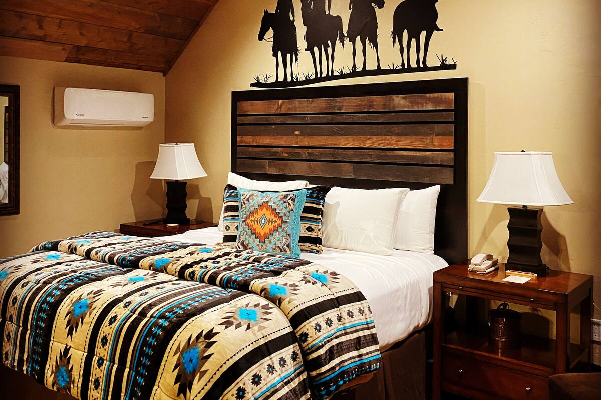A bedroom with a wooden ceiling, a bed with patterned bedding, two bedside lamps, and a wall decoration of horse riders.