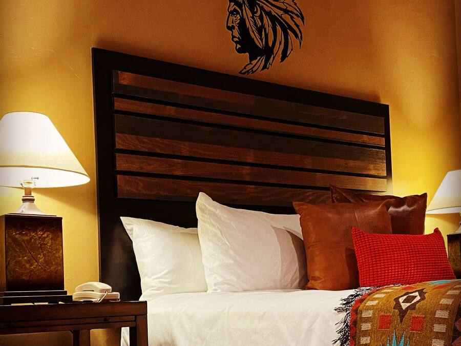Habitación de hotel con cabecera de madera, cama con ropa de cama blanca y marrón, un cojín decorativo rojo y una obra de arte tribal colgada en la pared.
