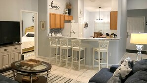 A modern apartment with a kitchen, dining area, and living room. It includes a TV, a sofa, and a small dining table.