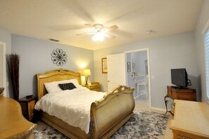 A bedroom with a wooden bed, a ceiling fan, a bedside table with a lamp, a television on a stand, and a door leading to another room. - Orlando BeachHouse ❤️ Expanded pool & HotTub Recent renos, close to Disney! (Davenport)
