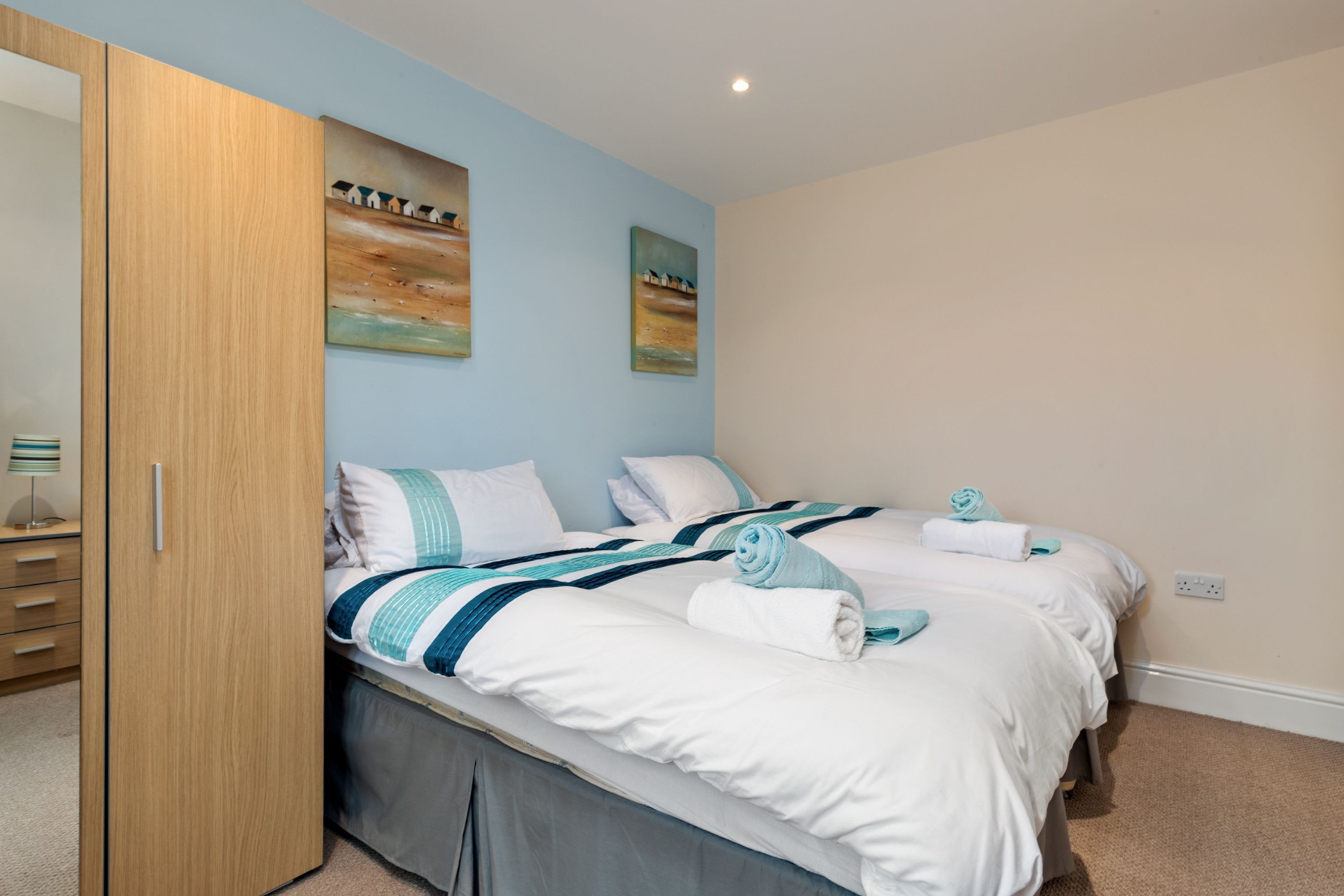 A bedroom with two beds, a wooden wardrobe, and two landscape paintings on the wall.