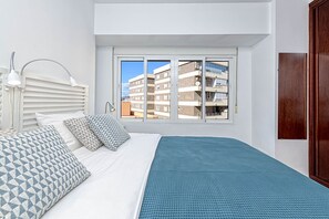 Une chambre à coucher avec un lit, des coussins à motifs, une lampe de chevet, une fenêtre donnant sur des immeubles et une porte en bois.