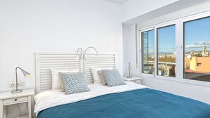 Un dormitorio con cama, dos lámparas de noche, una ventana y vistas a la ciudad, además de un cabecero blanco.