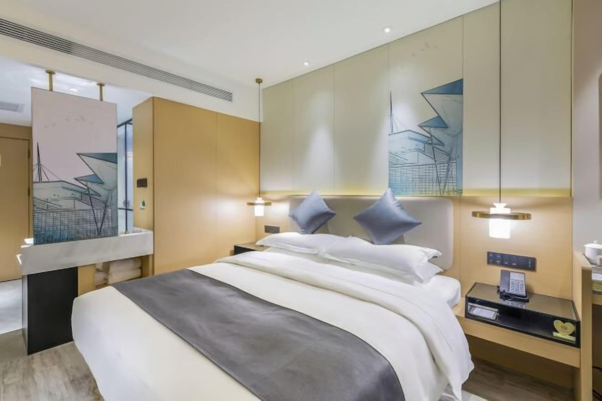 A modern hotel room with a large bed, bedside tables, a mirror, and abstract wall art.