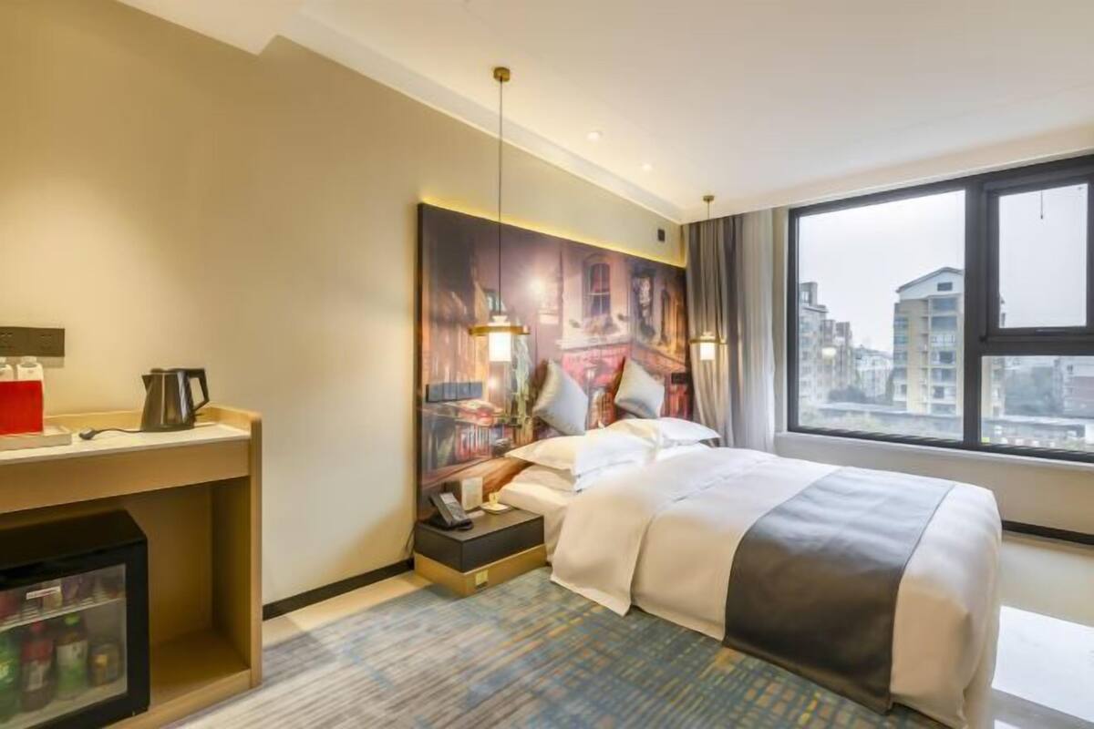 Habitación de hotel con una cama grande, un escritorio con tetera, un refrigerador y vistas a la ciudad por la ventana.