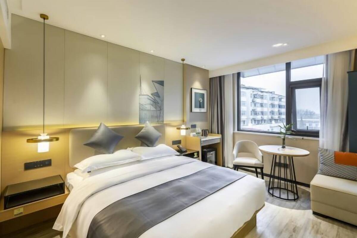 Habitación de hotel con cama, mesita de noche, lámpara, silla y vista a edificios a través del ventanal.