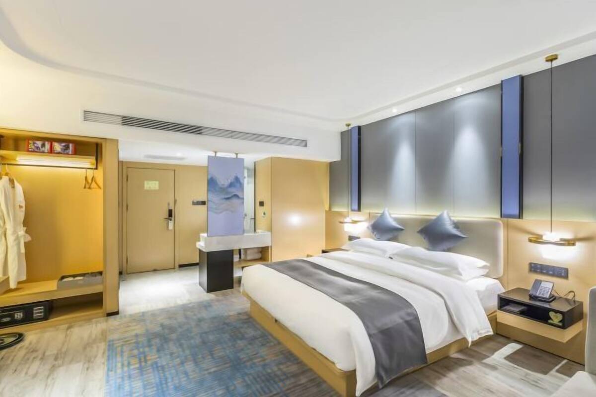 Una habitación de hotel moderna con una cama grande, una mesita de noche con lámpara, un armario y un baño con espejo y toallero.