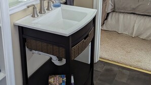 A bathroom with a large mirror, a white sink, and a black vanity. There is a rug on the floor and a bed in the adjacent room.