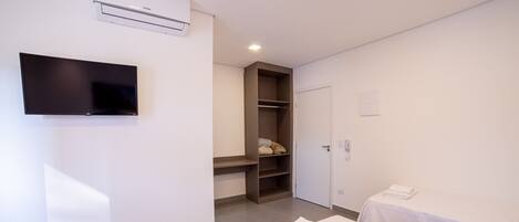 A hotel room with a flat-screen TV, a wardrobe, a bed with a white bedspread, and a small table with a folded towel.