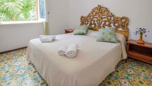 Una cama bien hecha con un tablero decorativo, dos almohadas y toallas enrolladas sobre un suelo estampado.