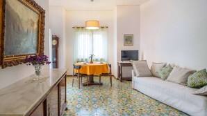 Una sala de estar con sofá, una mesa de comedor con sillas, televisor, reloj y un florero con flores.