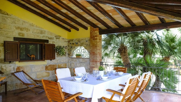 A covered outdoor dining area with a stone wall, wooden ceiling beams, and a view of palm trees. - Villa Virna with garden and pool - My Rental Homes (Cinisi)