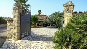 A stone gate with a metal fence, a paved courtyard, and a building with a tiled roof. - Villa Virna with garden and pool - My Rental Homes (Cinisi)