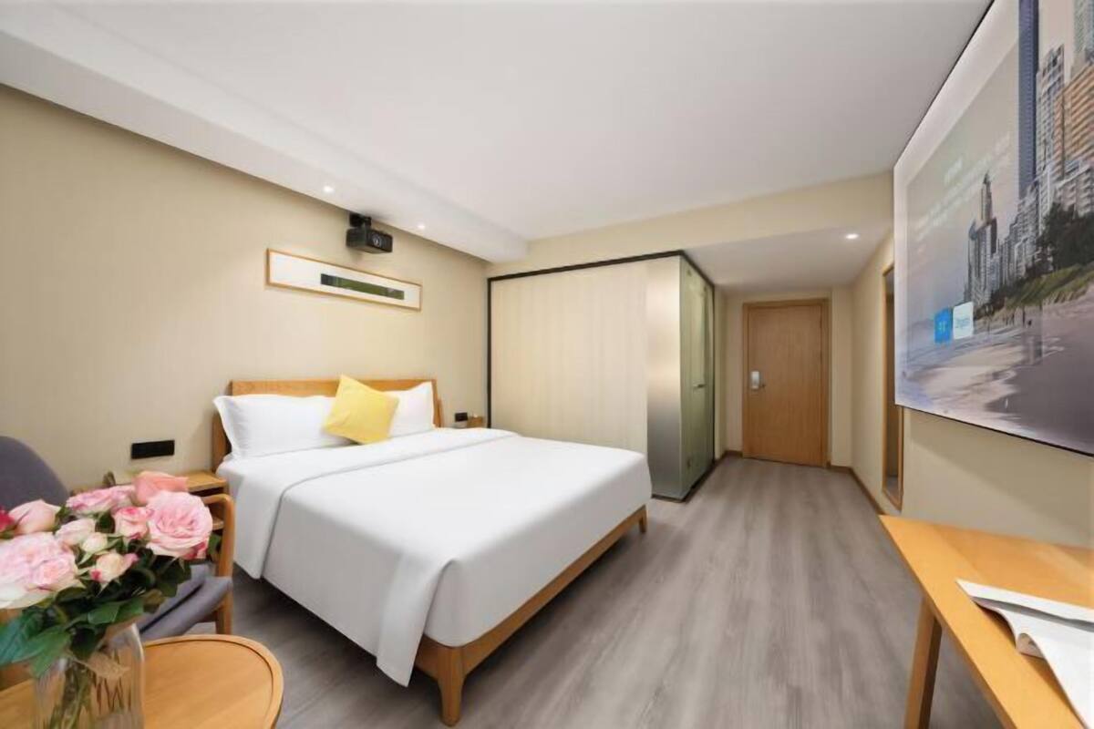 A hotel room with a bed, a desk, a chair, a vase with flowers, and a large window.