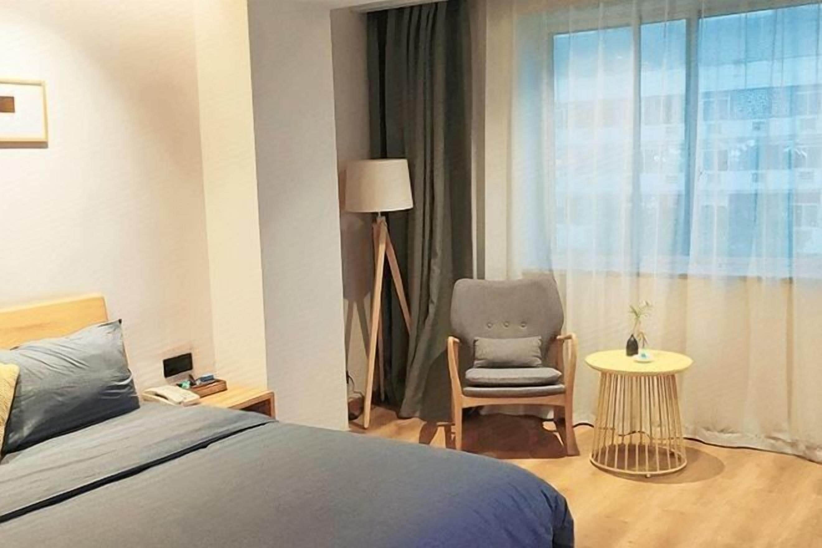Hotelkamer met een bed, een stoel, een tafeltje, een lamp en een raam met gordijnen.