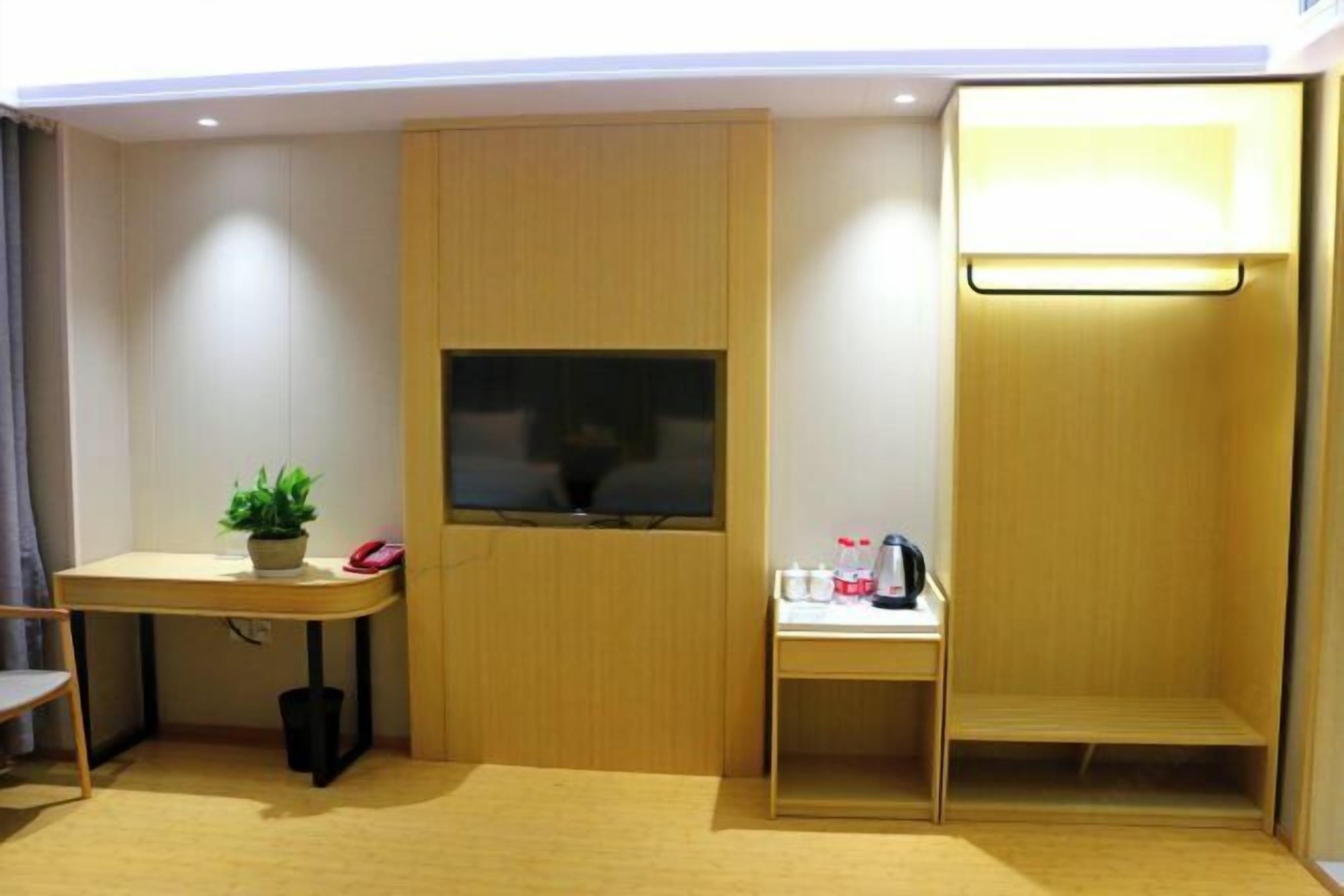 A compact room with a wooden desk, a small table with a plant, a trash bin, and a shelf with a kettle and cups.