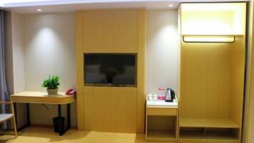 A compact room with a wooden desk, a small table with a plant, a trash bin, and a shelf with a kettle and cups.