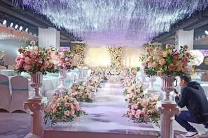 An indoor event space with a central aisle flanked by columns adorned with floral arrangements, leading to a stage with a backdrop. - Hanxiang International Hotel (Feidong)