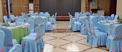 A banquet hall with round tables and chairs, a stage, and a projector screen.