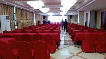 Un salón de banquetes con filas de mesas cubiertas con manteles rojos y sillas, suelo de baldosas y un largo pasillo que conduce a una puerta.