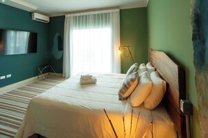 Habitación de hotel con una cama grande, un televisor de pantalla plana montado en la pared, una ventana con cortinas translúcidas y una lámpara de noche.
