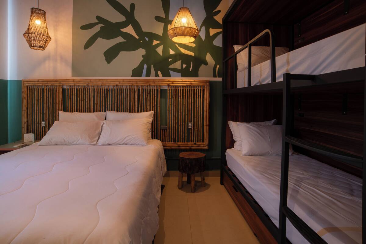 A hotel room with a large bed, a smaller bed, and a bunk bed. There are two hanging lamps and a small round table.