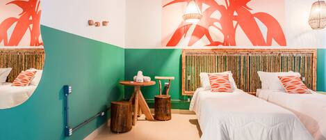 Habitación de hotel con dos camas, una mesita con una silla, dispensador de toallas en la pared y un cuadro abstracto grande.