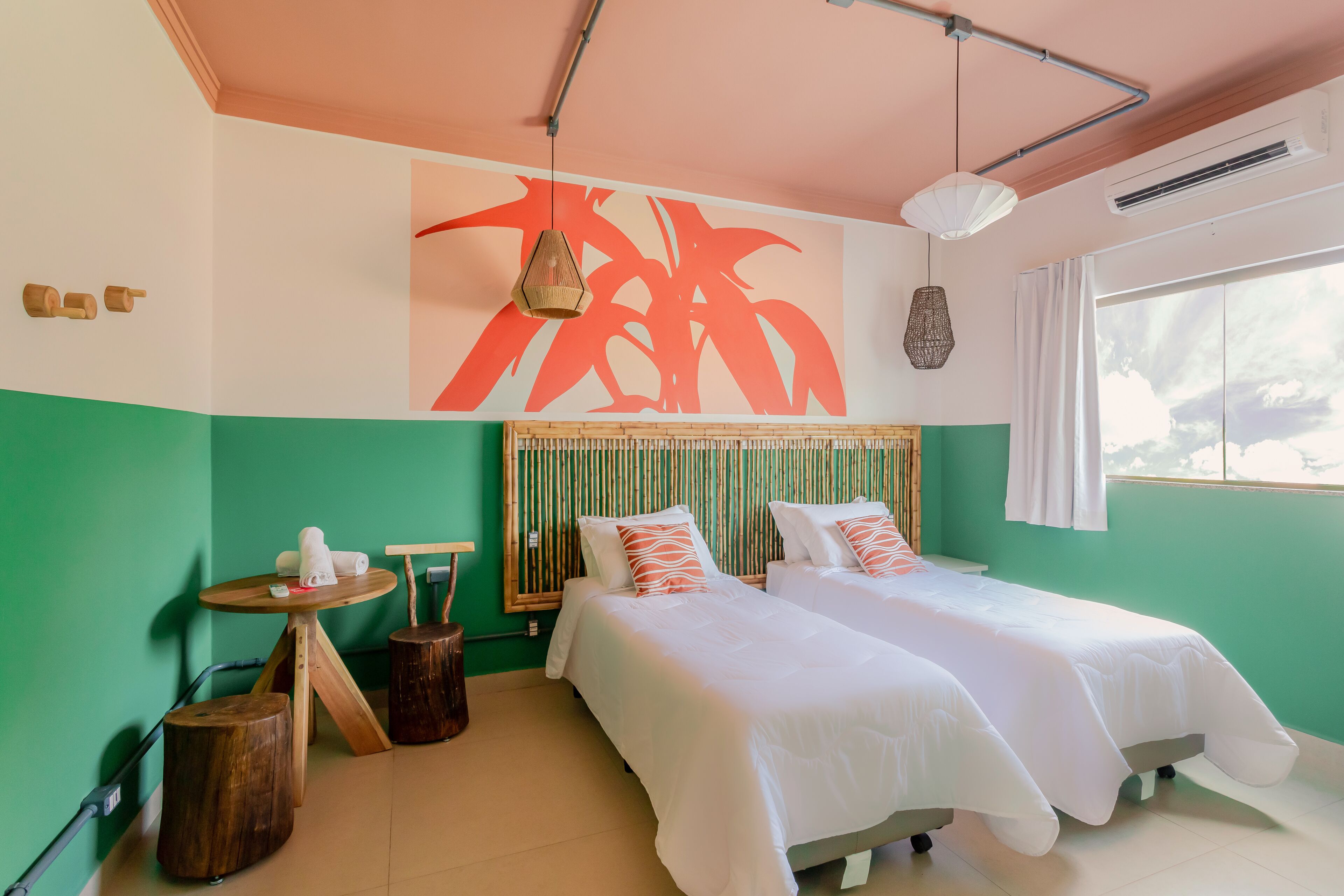 A room with two beds, a round wooden table with a towel, a hanging light, and a large abstract wall art piece.