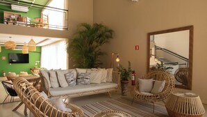 Amplia sala de estar con techo alto, un sofá grande, mobiliario de mimbre, una planta en maceta y una escalera visible a través de un panel de vidrio.