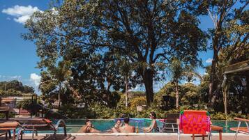 Zona junto a la piscina con sillones de descanso y una mesa, rodeada de árboles y un cielo despejado.