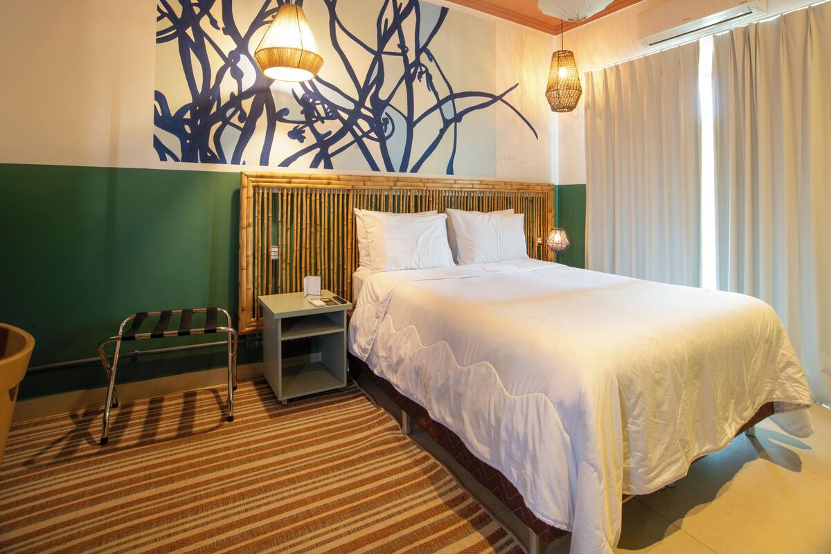 A hotel room with a bed, a nightstand, a chair, a wall art piece, and a window with curtains.