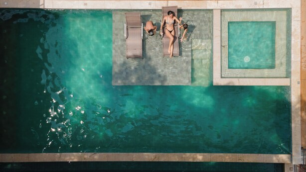 Vista aérea de una piscina con un borde de baldosas, sillas de descanso y una sección de la piscina en un tono azul diferente.