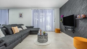 Una sala de estar moderna con un sofá modular, una mesa redonda de centro, un televisor de pantalla plana montado en la pared y un pequeño taburete naranja.