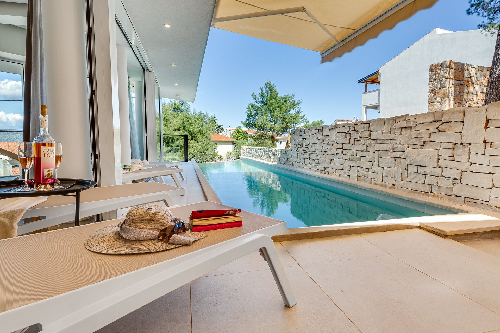A poolside area with lounge chairs, a table with a bottle and glasses, and a stone wall.