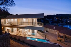 Een modern huis met twee verdiepingen, een glazen gevel, een zwembad en uitzicht op een stad en een waterpartij in de schemering.