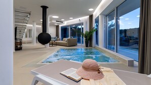 A modern indoor swimming pool with a view of the outdoors, a staircase, and a cozy seating area.