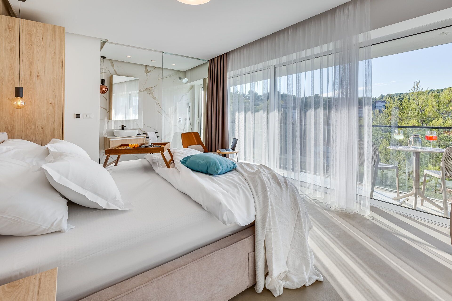 Una habitación de hotel moderna con una cama grande, cabecera de madera, una mesita con una computadora portátil, y un balcón con mesa y sillas.