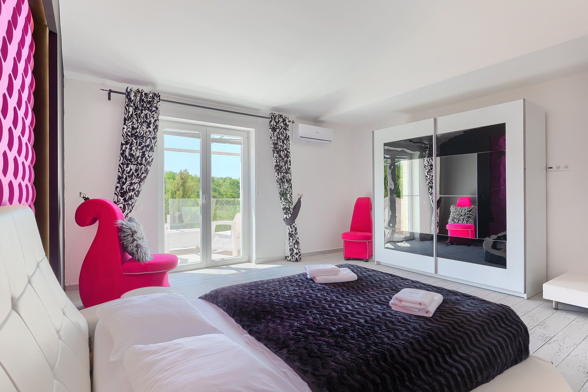 Ein Schlafzimmer mit einem großen Bett, einer Schiebetür zu einem Balkon, einem Spiegelkleiderschrank und einem rosa Sessel.