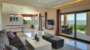 Een moderne woonkamer met een pooltafel, een bank, een salontafel en een televisie. Er zijn grote ramen met uitzicht op de omgeving.