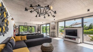 A modern living room with a large sofa, a round coffee table, and a flat-screen TV. There is a kitchen area with a dining table and chairs, and a large window offering a view of the outdoors.