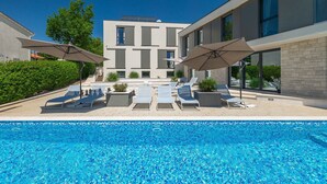A modern pool area with lounge chairs and umbrellas, surrounded by a building with large windows and a stone facade.