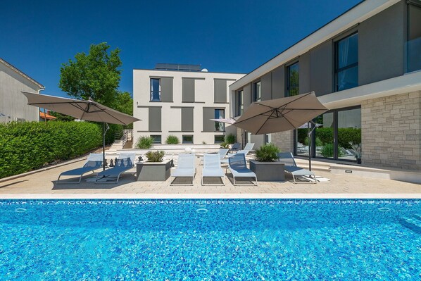 Ein moderner Poolbereich mit Liegestühlen und Sonnenschirmen, umgeben von einem Gebäude mit großen Fenstern und einer Stein-Fassade.