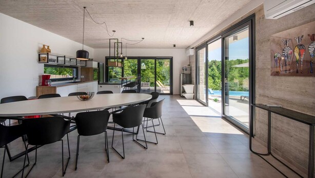 A modern dining area with a large table and chairs, a kitchen with a bread sign, and a view of a swimming pool through large glass doors.