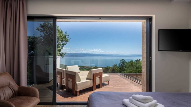 Une chambre d’hôtel moderne dotée d’une grande porte-fenêtre coulissante donnant sur un balcon avec une vue imprenable sur un lac et des montagnes. On y trouve un lit avec une serviette soigneusement pliée et un fauteuil en cuir.